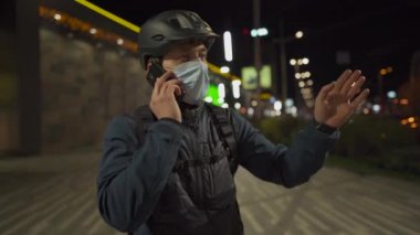 Tıbbi maske ve kask takan bisikletçi, karantina koronavirüsü sırasında şehre bisiklet teslimatına gitmeden önce bir sonraki müşteriyle telefonda konuşur. Bisikletli kurye, banliyö