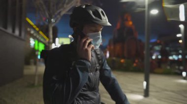 Kasklı ve koruyucu maskeli bisikletli bir bisikletçi işten eve dönerken gece telefonda konuşuyor. Bisikletli teslimatçı, teslimat paketleri, koronavirüs karantinası sırasında telefon görüşmesi.