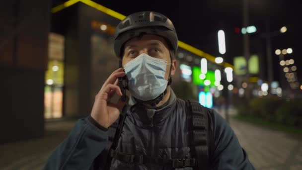 Cyclist in helmet and protective mask commuter with bicycle on way home from work talking on phone at night city. Deliveryman on bike, delivery parcels, make phone call during coronavirus quarantine — Stock Video