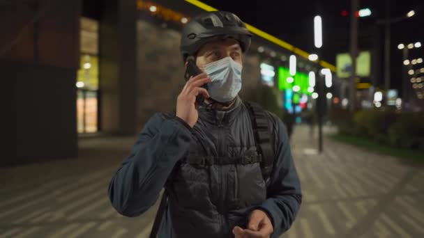 자전거를 타고 퇴근하는 길에 헬멧 과 보호용 마스크를 착용 한 자전거 여행자가 밤 도시에서 전화 통화를 하고 있다. 자전거를 타고, 배달 소포를 배달하고, 코로나 바이러스 검역중에 전화를 걸고 — 비디오