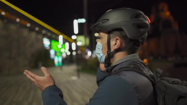 Cyclist in helmet and protective mask commuter with bicycle on way home from work talking on phone at night city. Deliveryman on bike, delivery parcels, make phone call during coronavirus quarantine — Stock Video