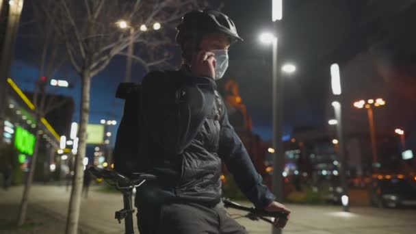 Homem de entrega masculino com bicicleta usando capacete e máscara protetora contra vírus vívido e poluição, entrega parcelas na cidade à noite em bicicleta, fazendo telefonema para o cliente durante a quarentena do coronavírus — Vídeo de Stock