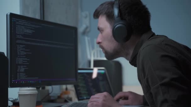 自宅からヘッドフォンでノートパソコンの後ろに動作しながらハンサムな男性ソフトウェア開発者のプログラミングコード。オフィスのコンピュータで働くヘッドフォンのITエンジニア。フリーランスの仕事、社会的距離 — ストック動画