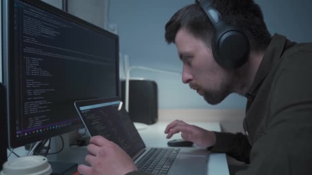 Jóképű férfi szoftverfejlesztő programozási kódok, miközben a laptop mögött dolgozik fejhallgatóval otthonról. Informatikai mérnök a fülhallgatóban, számítógépen dolgozik az irodában. Szabadúszó munkák, társadalmi távolság — Stock videók