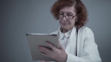 Gözlüklü ve tıbbi önlüklü orta yaşlı beyaz kadın doktor dijital tabletle çalışıyor ve klinikteki danışmanlık yapan hastaya yönelik ilaçlar hakkında bilgi topluyor. İlaç ve sağlık hizmetleri