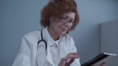 Gözlüklü ve tıbbi önlüklü orta yaşlı beyaz kadın doktor dijital tabletle çalışıyor ve klinikteki danışmanlık yapan hastaya yönelik ilaçlar hakkında bilgi topluyor. İlaç ve sağlık hizmetleri