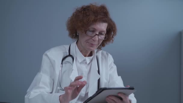 中世の白人女性医師が眼鏡や医療用ガウンを着用してデジタルタブレットを使用しており、診療所で患者を診察するための医薬品に関する情報を研究している。医学・医療 — ストック動画