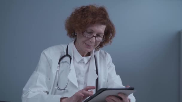 Geneeskunde en gezondheidszorg. Portret gelukkig senior vrouwelijke arts met patiëntenkaart op digitale tablet werken aan tafel in het ziekenhuis kantoor. Telegeneeskunde app op afstand in het ziekenhuis werkt online — Stockvideo