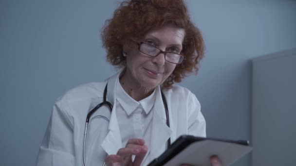 Concepto de medicina y salud. Retrato feliz médico senior femenino sosteniendo la carta del paciente en la tableta digital que trabaja en la mesa en la oficina del hospital. Aplicación de telemedicina remota en el hospital trabajando en línea — Vídeos de Stock