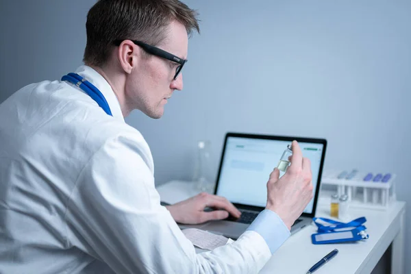 Young caucasian doctor working at table in hospital laboratory examining an ampoule of medicine and entering data into computer. New vaccine against coronavirus. Scientist changing future covid 19 — Stock Photo, Image
