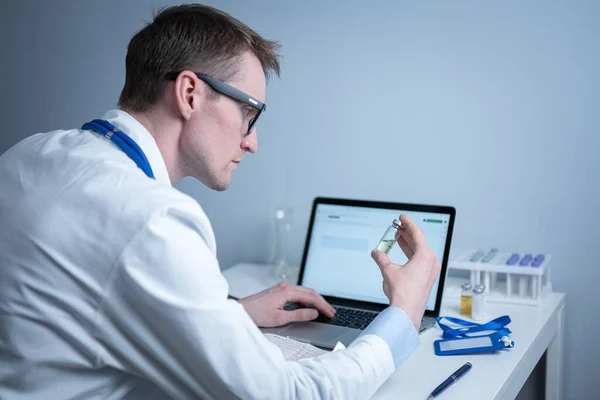 Young caucasian doctor working at table in hospital laboratory examining an ampoule of medicine and entering data into computer. New vaccine against coronavirus. Scientist changing future covid 19 — Stock Photo, Image