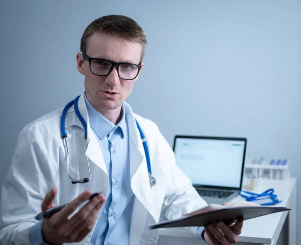 Young male doctor consults patient in hospital office, holding medical examinations in hands. General practitioner in white medical coat looks into camera and makes an appointment with patient online