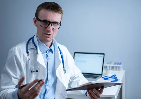 Giovane medico maschio consulta il paziente nello studio ospedaliero, tenendo esami medici in mano. Medico generico in camice medico bianco guarda nella fotocamera e prende un appuntamento con il paziente online — Foto Stock