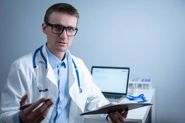 Young male doctor consults patient in hospital office, holding medical examinations in hands. General practitioner in white medical coat looks into camera and makes an appointment with patient online