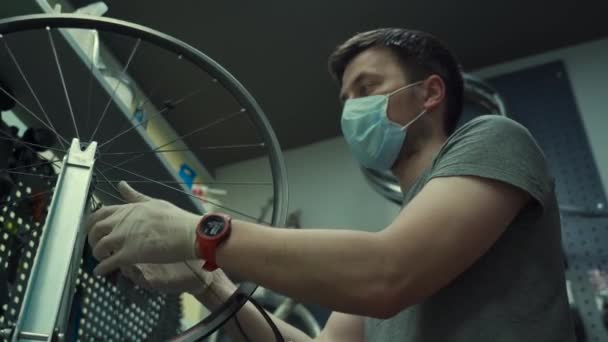 Bicycle shop repairman works in bicycle service and repair workshop during coronavirus quarantine wearing face shield and gloves, new norm. Mechanic repairing bicycle wheel wearing medical mask — Stock Video
