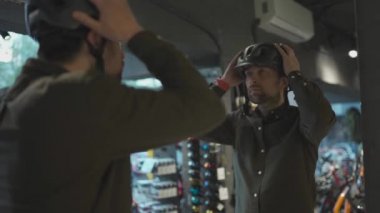 Genç adam bisiklet mağazasına geldi. Miğferi ölçüyor. Erkek, spor malzemeleri mağazasında kask seçer. Yeni bir kask satın almak. Bisiklet kasklı müşteri aynanın yanında deniyordu.