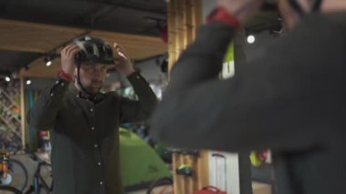 Spor malzemeleri mağazasında aynanın yanında siyah bisiklet kaskı deneyen beyaz bir adam. Erkek alıcı bisiklet sürmek için kaskı seçer. Bisiklet dükkanından alışveriş. Kişi, kask kayışını ayarlar.