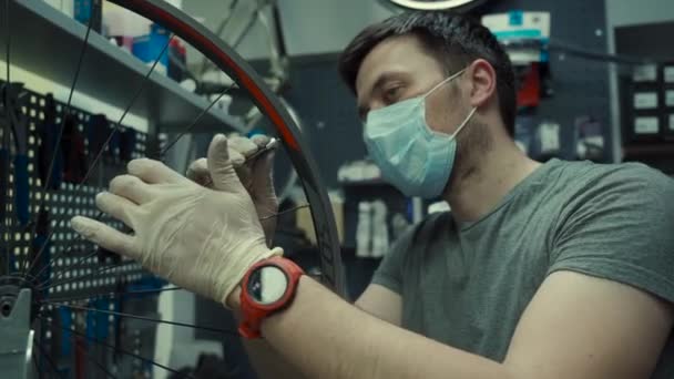 Mekaniker reparerar cykel i verkstad bär covid 19 mask. Arbetare av cykelaffär fixar cykel under coronavirus karantän i skyddsmask. Underhållscykel för reparatörer. Sportbutikskoncept — Stockvideo