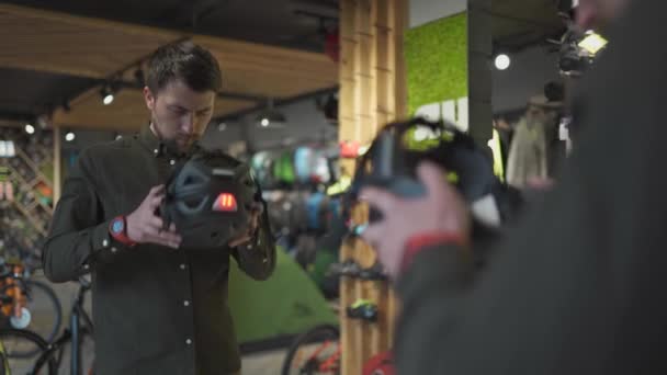 Hombre examinando cascos de bicicleta en la tienda de deportes mirándose en el espejo. Probando casco deportivo nuevo en la tienda de bicicletas. Hombre que se pone el casco de bicicleta en la tienda. Cliente en tienda de bicicletas probándose el casco de bicicleta — Vídeos de Stock