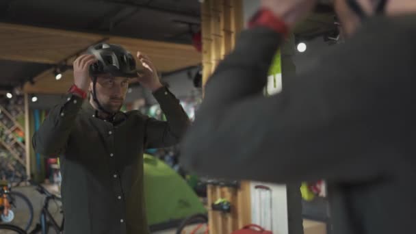 Hombre caucásico probándose el casco de bicicleta negro cerca del espejo en la tienda de artículos deportivos. Hombre comprador elige casco de seguridad para el ciclismo. Compras en la tienda de bicicletas. Persona ajusta su correa de casco deportivo — Vídeo de stock