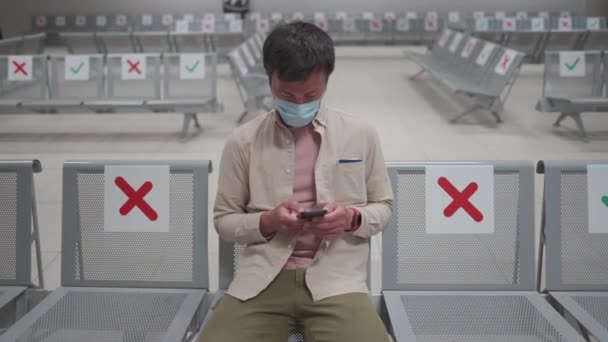 Traveler sits at international airport of Cyprus, Paphos during COVID-19 crisis, sits in terminal on seat with social distance markings wearing mask. Using phone while waiting to board airline flight — Stock Video