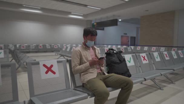 Kıbrıs uluslararası havaalanında oturan gezgin, COVID-19 krizi sırasında Paphos, maske takan sosyal mesafe işaretleriyle terminalde oturuyor. Uçağa binmek için telefon kullanmak. — Stok video
