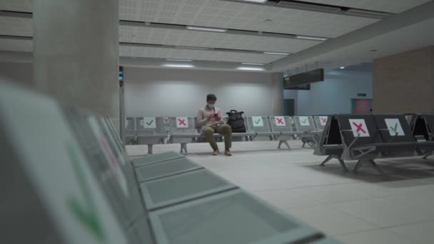 旅行者は、 COVID-19危機の間にパフォスのキプロスの国際空港に座って、マスクを身に着けている社会的距離マーキングと座席のターミナルに座っています。飛行機に乗るのを待っている間に電話を使う — ストック動画