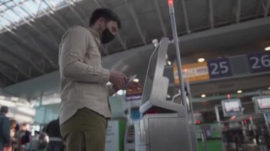 Koruyucu maskeli bir adam havaalanında kendini kontrol gişesini kullanıyor. Havaalanında giriş kartı alan bir erkek var. Sosyal mesafe, karantina covid 19 sırasında terminalde otomatik kontrol bagajı.