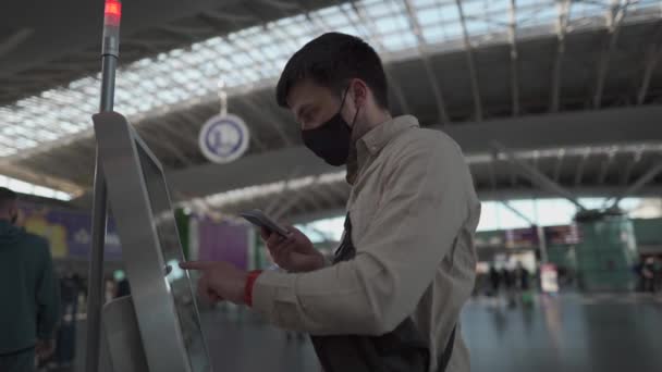 Человек в маске садится на самолет в аэропорту. Трейлер на сенсорном экране станции саморегистрации в терминале. Регистрация рейсов, вспышка коронавируса. Машина самообслуживания в зоне отправления — стоковое видео