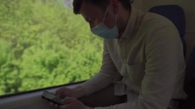Yeni toplu taşıma standardı, trenle seyahat eden kişi güvenli mesafede oturur ve maske takarak telefon kullanır. Adam pencerenin yanındaki trende akıllı telefon kullanıyor. Covid 19 'un yayılmasını önlemek için maskeli gezgin