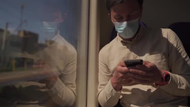 Maskovaný cestující používají telefon ve veřejné dopravě, když sedí u okna ve vlaku. Dívá se na obrazovku, používá mobilní internet. Koncept je nová norma a sociální vzdálenost při cestování po železnici — Stock video