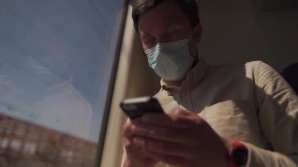 Nuova norma dei trasporti pubblici, persona viaggia in treno si siede a distanza di sicurezza e utilizza telefono indossando maschera. L'uomo usa smartphone all'interno del treno vicino alla finestra. Viaggiatore in maschera per prevenire la diffusione di Covid 19 — Video Stock