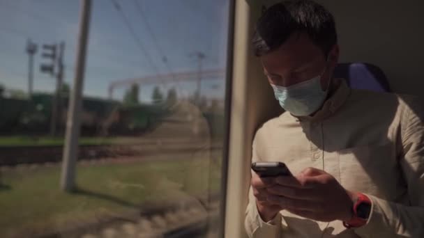 Masked passenger use phone in public transport while sitting by window in train carriage. Looks at screen, uses mobile Internet. Concept is new norm and social distance while traveling on railway — Stock Video