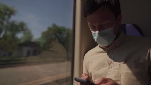 Nuova norma dei trasporti pubblici, persona viaggia in treno si siede a distanza di sicurezza e utilizza telefono indossando maschera. L'uomo usa smartphone all'interno del treno vicino alla finestra. Viaggiatore in maschera per prevenire la diffusione di Covid 19 — Video Stock