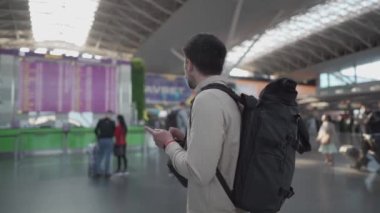 Sırt çantalı maskeli adam tekrar seyahat etmekten mutlu, uçuş bilgi panosunun yanındaki akıllı telefonu kullanıyor. Koruyucu maskeli bir erkek yolcu uçuş programını izliyor ve bekliyor.
