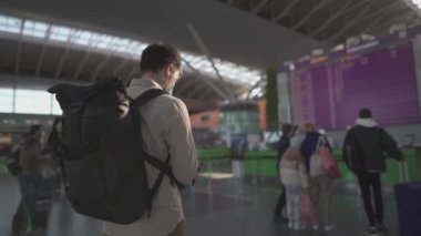Sırt çantalı maskeli adam tekrar seyahat etmekten mutlu, uçuş bilgi panosunun yanındaki akıllı telefonu kullanıyor. Koruyucu maskeli bir erkek yolcu uçuş programını izliyor ve bekliyor.