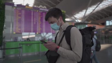 Sırt çantalı maskeli adam havaalanı terminalinde, uçuş çizelgesinde bekliyor. Maskeli bir erkek elektronik çizelgeye yakın telefon kullanıyor, kontrol gişesinin açılmasını bekliyor