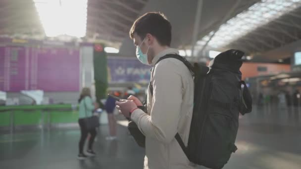 Zamaskowany człowiek z plecakiem stojący przy planach linii lotniczych na lotnisku, covid-19, nowy normalny. Mężczyzna w masce używać telefonu w pobliżu elektronicznego harmonogramu, czekając na otwarcie kasy odpraw — Wideo stockowe