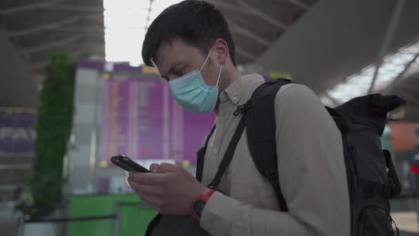 Maskovaný cestující s batohem na letišti se dívá na letový řád a používat telefon, nové pravidlo a předpisy pro cestování letadlem během covid19. Backpacker v masce v odbavovací oblasti elektronický jízdní řád — Stock video