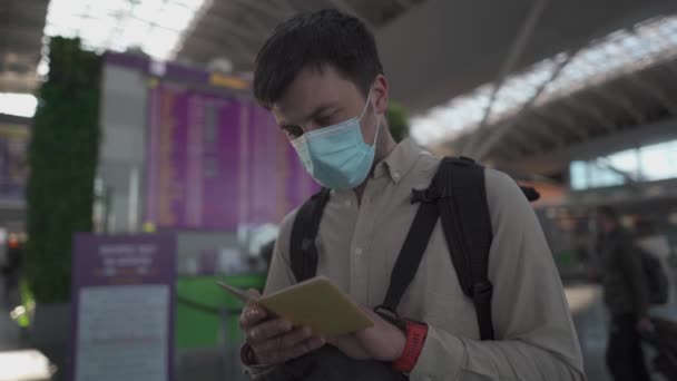 戴面具的背包客高兴地再次旅行，使用智能手机接近飞行信息板。持防护面具的男性旅客在登机时间监视器上观看和等待航班时刻表 — 图库视频影像