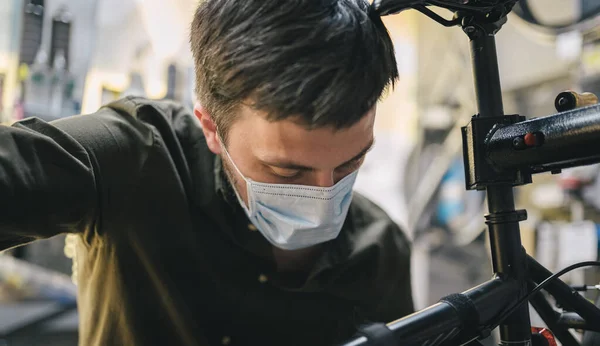 Opravář motocyklů pracuje v cyklistické servisní a opravárenské dílně během karantény koronaviru s obličejovým štítem a rukavicemi, nová norma. Mechanik opravy kolo nosí lékařskou masku — Stock fotografie