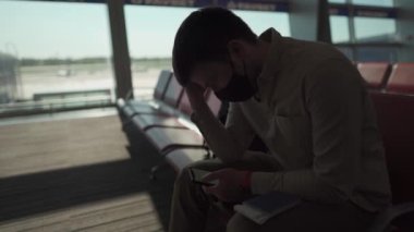 Koruyucu maskeli bir adam uçağa binmek için bekliyor, internette sörf yapıyor, Ukrayna 'daki Boryspil terminalinde telefon kullanıyor, hava güneşli ve sosyal mesafeli.
