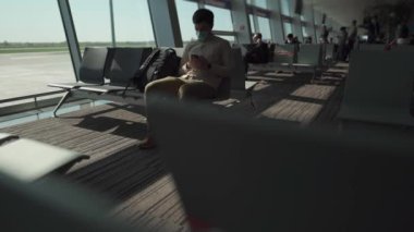 Hava yolculuğu için yeni normal bir kavram. Havaalanında sosyal mesafeler. Akıllı telefonunu kullanarak kovid 19 maskesi takan bir adam uçağa binmeden önce lobide oturur. Koronavirüs sırasında tek başına seyahat etmek.