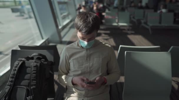 Uomo mascherato si siede all'aeroporto sul sedile distanza sociale dal finestrino prima di salire sull'aereo e utilizzare il telefono per noia. Ritardo del volo. Cancellazione dei voli a causa di quarantena e isolamento covid 19 — Video Stock