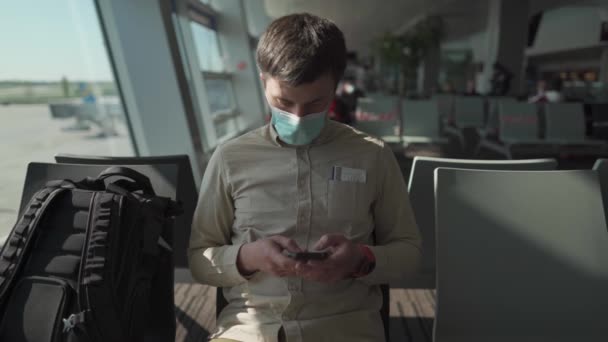 新的正常的航空旅行概念。机场的社交场合。在登机前，一个戴着系数19的面具，用智能手机的男人坐在大厅里。在验尸过程中独自旅行 — 图库视频影像