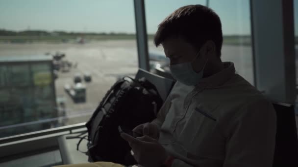 Man passagerare med ryggsäck i skyddsmask väntar på att gå ombord på ett flygplan, surfa på Internet, med hjälp av telefon i terminal Boryspil flygplats i Ukraina, soligt väder genom fönster på sociala avståndsplatser — Stockvideo