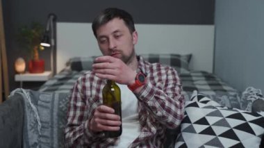 Genç yaşta alkol bağımlılığının teması. Kafkasyalı sarhoş adam bir şişe şarapla konuşuyor ve evde kanepede alkol içiyor. Alkolizm ve umutsuzluk. Kötü alışkanlık. Sarhoş olmak. Tek başına alkol içiyor.