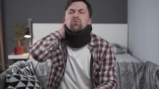 Guy trägt Nackenstütze, um den Nacken zu Hause auf der Couch zu stützen. Kaukasischer Mann mit Nackenverletzung nutzt Kragenstütze, um Schmerzen zu lindern. Männchen in Halswirbelsäule lindert Verspannungen und Schmerzen in den Muskeln der Wirbelsäule — Stockvideo
