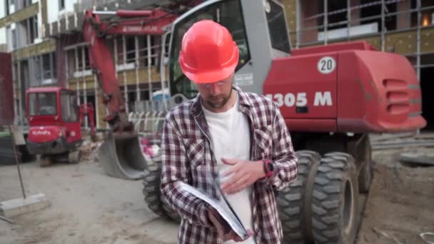 Hombre caucásico ingeniero arquitecto que sostiene la carpeta con papeles en la excavadora de fondo, maquinaria de construcción en el sitio de construcción. Constructor mirando el plan de negocios, documentos arquitectónicos — Vídeo de stock