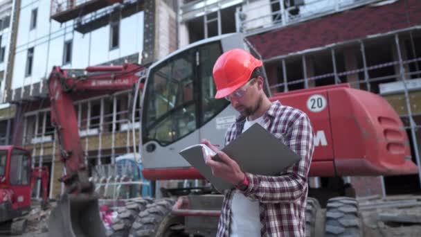 Manajer konstruksi muda memeriksa dokumentasi teknis saat berada di lokasi konstruksi dekat traktor atau ekskavator alat konstruksi berat. Mandor memeriksa laporan — Stok Video